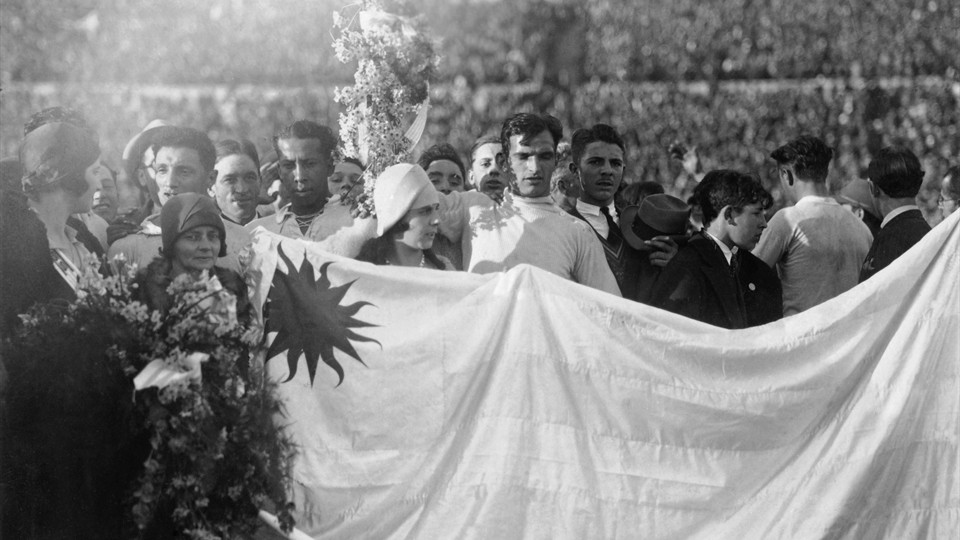 جام جهانی 1930|دروازه بان اروگوئه، انریکه بالستروس (وسط) در حالی که اعضای تیم، هواداران و مقامات پشت پرچم ملی اروگوئه به صف ایستاده اند دسته گلی را در هوا بلند کرده. این عکس پس از قهرمانی اروگوئه در فینال جام جهانی 1930 برابر آرژانتین، در ورزشگاه سنتناریو در مونته ویدئو گرفته شده است. 30 جولای 1930. اروگوئه 4-2 به پیروزی رسید.