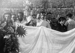 جام جهانی 1930|دروازه بان اروگوئه، انریکه بالستروس (وسط) در حالی که اعضای تیم، هواداران و مقامات پشت پرچم ملی اروگوئه به صف ایستاده اند دسته گلی را در هوا بلند کرده. این عکس پس از قهرمانی اروگوئه در فینال جام جهانی 1930 برابر آرژانتین، در ورزشگاه سنتناریو در مونته ویدئو گرفته شده است. 30 جولای 1930. اروگوئه 4-2 به پیروزی رسید.